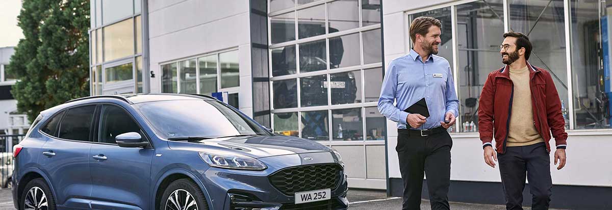 Zwei Männer unterhalten sich neben einem blauen Ford SUV vor einer Werkstatt.