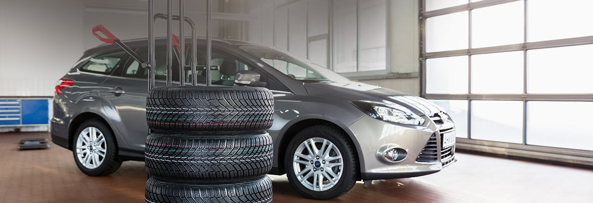 Ein graues Auto steht in einer Werkstatt neben einem Stapel Reifen und Werkzeug, helle Umgebung.