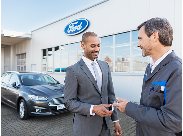 Ein Mann in einem Anzug erhält Schlüssel von einem Händler vor einem Ford-Autohaus.