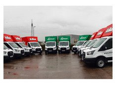 Mehrere Lieferwagen mit roten und grünen Aufbauten stehen auf einem Parkplatz. Bewölkter Himmel im Hintergrund.